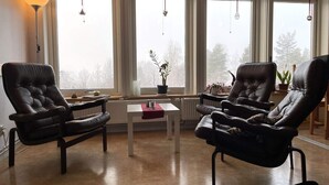 A living room with two leather armchairs, a small white table, and a floor lamp. There are three windows with curtains and hanging light fixtures. - 2 Person Holiday Park Home in Orsa (Orsa)