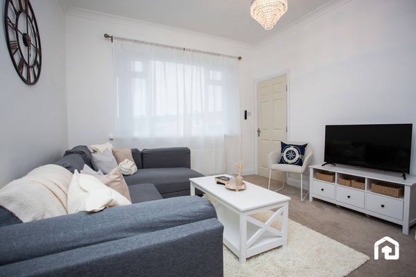 Relax in this bright, cozy living room featuring a comfy sectional sofa, soft neutral decor, and a flat-screen TV. The large window lets in plenty of natural light, creating a warm and inviting atmosphere.