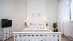 A bedroom with a white crib, two bedside tables with lamps, a TV, and framed pictures on the wall. - Vacant Nests - The Melrose | Beach | Free Parking (Northumberland)