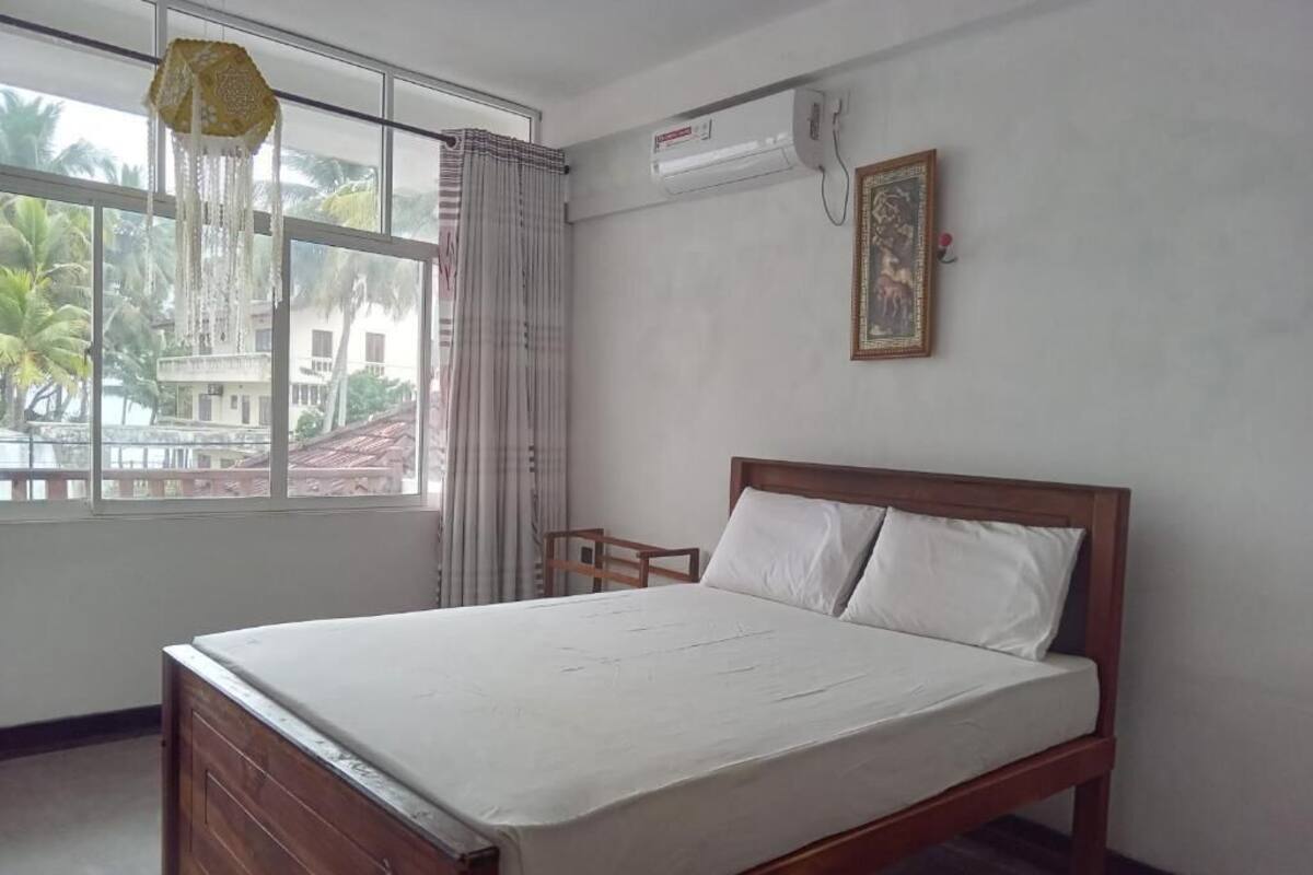A bedroom with a wooden bed, a window with curtains, an air conditioning unit, and a framed picture on the wall.
