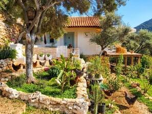A rustic house with a stone wall, a wooden deck, and chickens in the garden.
