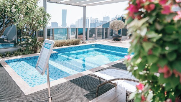 A rooftop swimming pool with a city skyline view, surrounded by greenery and lounge chairs. - Summer 1BR Sanctuary l Central l Balcony Canal View l Downtown (Dubai)