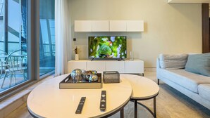 A modern living room with a large flat-screen TV, a sofa, a coffee table, and a dining area with chairs and a tray of items. - Summer 1BR Sanctuary l Central l Balcony Canal View l Downtown (Dubai)
