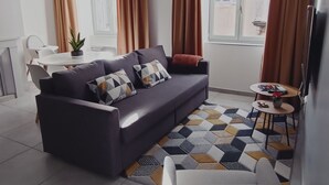 Una sala moderna con un sofá, una mesita y una silla. Se aprecia una ventana con cortinas, una lámpara en el techo y un suelo de baldosas.