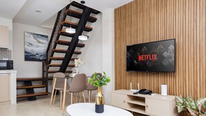 Moderni olohuone, jossa on portaikko, litteänäyttöinen televisio, jossa näkyy Netflix, ruokailutila ja viherkasvi.