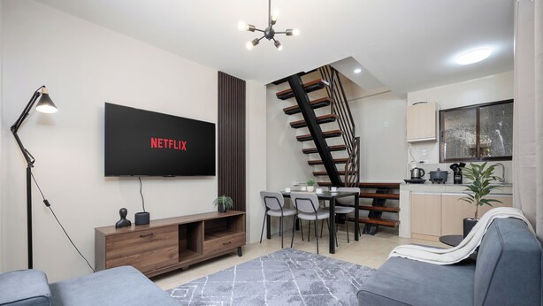 Ein modernes Wohnzimmer mit Flachbildfernseher, grauen Sofas, einer Holzeinbauküche und einer Treppe, die in einen Küchenbereich führt.