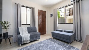 Ein modernes Wohnzimmer mit zwei grauen Sofas, einem gemusterten Teppich, einer Holztür und großen Fenstern mit Vorhängen.