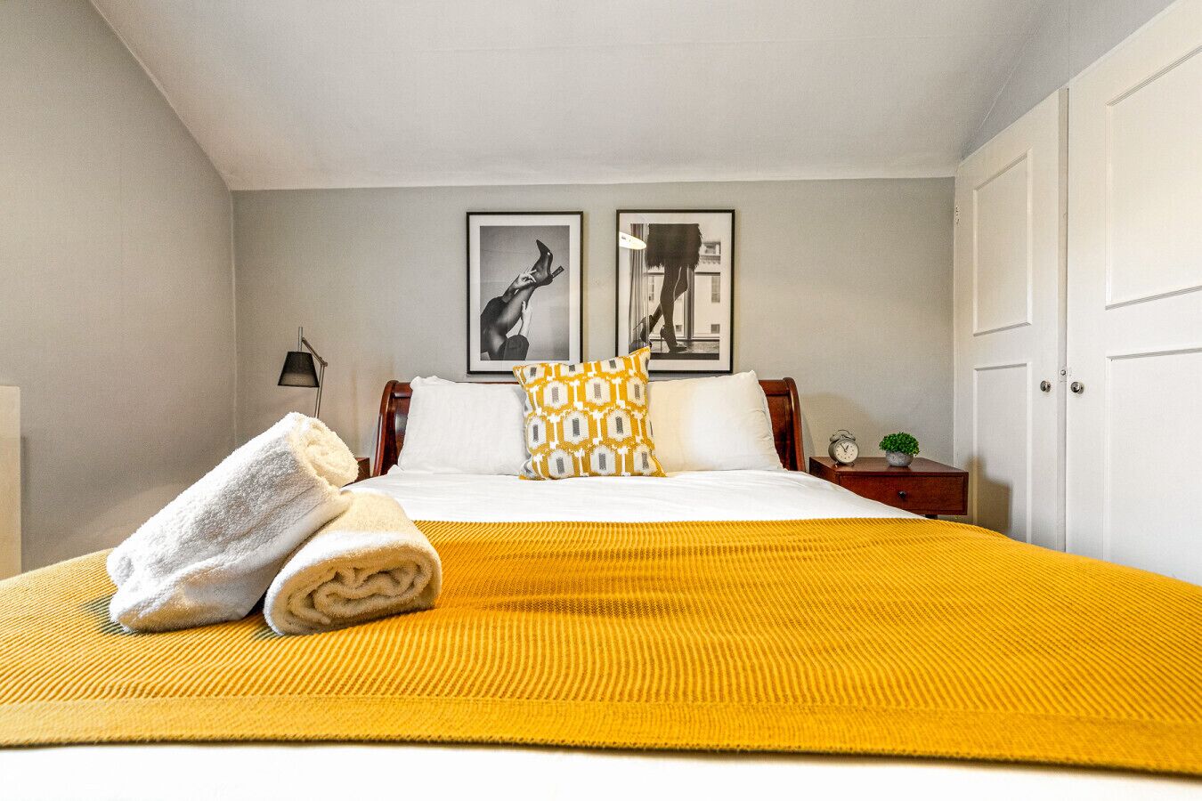 A neatly made bed with a yellow blanket, a white towel, and patterned pillows. There are two framed pictures on the wall, a small bedside table with a clock and a plant, and a white door.