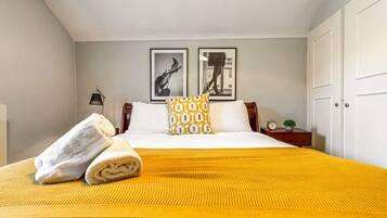 A neatly made bed with a yellow blanket, a white towel, and patterned pillows. There are two framed pictures on the wall, a small bedside table with a clock and a plant, and a white door.