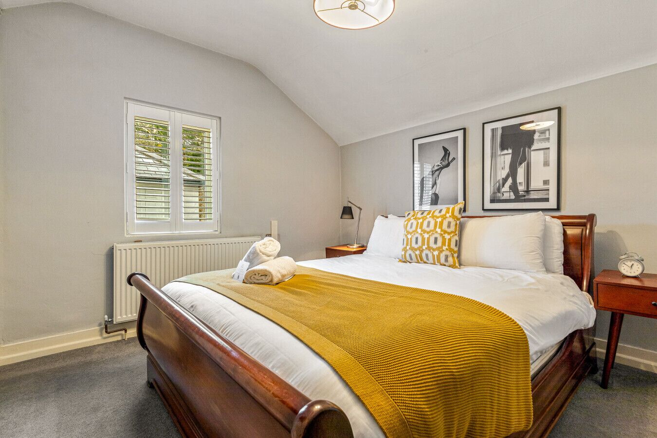Une chambre avec un lit en bois, un plaid jaune, un oreiller blanc, une table de chevet avec une lampe et un tableau encadré accroché au mur.