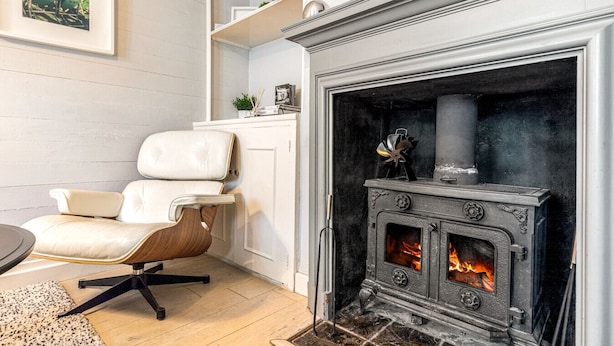 A modern living room with a white leather armchair, a fireplace with a vintage stove, and a wooden floor. - Mills Cottage - Sleeps 4 (Henley-On-Thames)