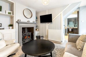 Una sala de estar con chimenea, un televisor de pantalla plana montado en la pared y una cocina visible a través de un umbral.