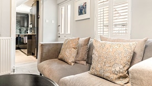 A living room with a beige sofa, patterned cushions, a small table, and a kitchen in the background. - Mills Cottage - Sleeps 4 (Henley-On-Thames)