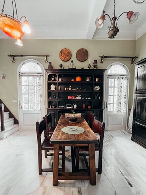 Un coin repas avec une table en bois, des chaises et un lustre. On y trouve une bibliothèque remplie de divers objets, ainsi qu’une porte dotée d’une fenêtre.