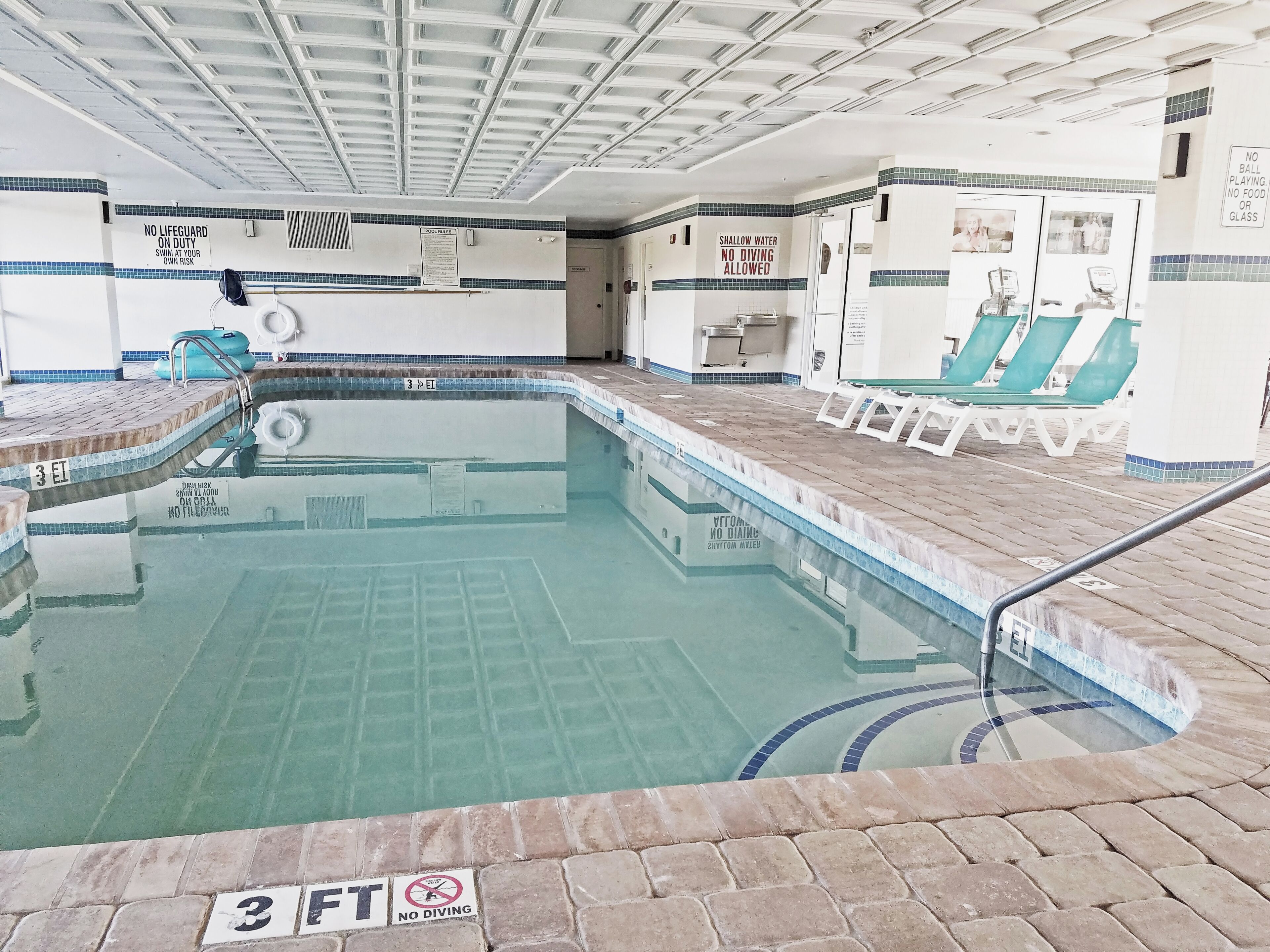 Une piscine intérieure avec un sol carrelé, un poste de surveillance pour le sauveteur et un panneau indiquant qu’il est interdit de plonger.
