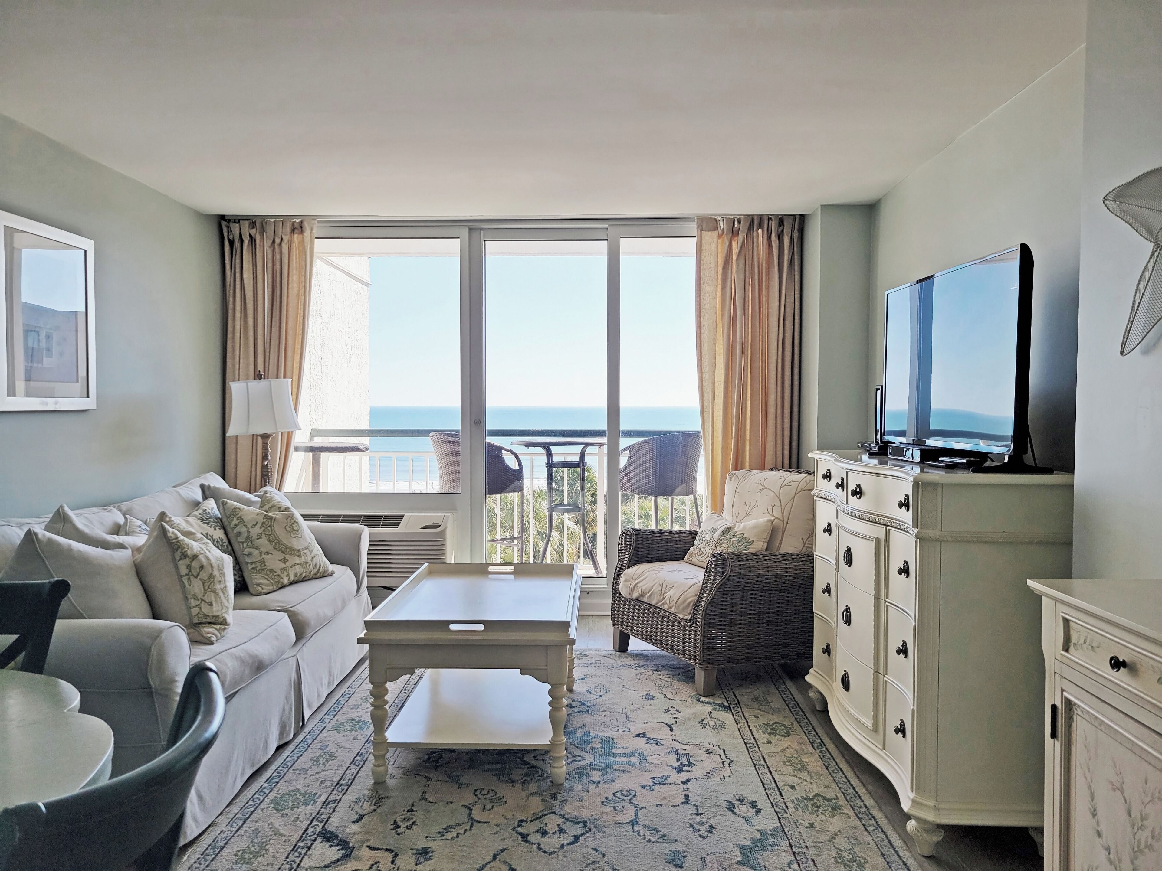 A hotel room with a sofa, a coffee table, a TV, a dining table, and a view of the sea.