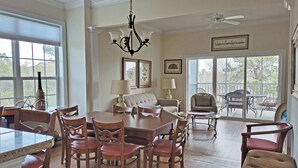 A spacious dining area with a wooden table and chairs, a chandelier, and a balcony with outdoor furniture. - Seaside Inn Condo, Screen Porch, Quiet Setting, Pool Plaza, Resort Beach Access (Pawleys Island)