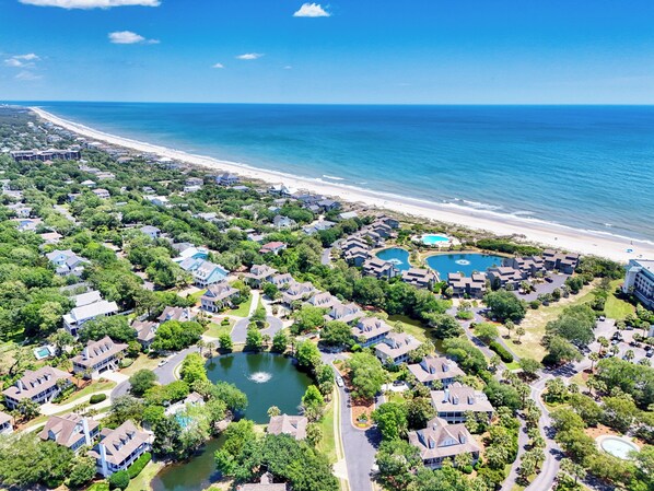 An aerial view of a coastal resort with multiple buildings, a central pond, and a beach.