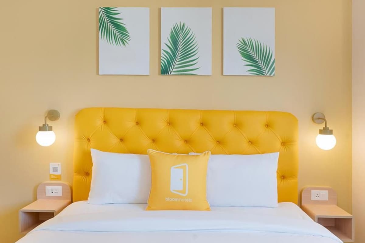 A hotel room with a yellow tufted headboard, white bedding, and three leaf-patterned wall art pieces.