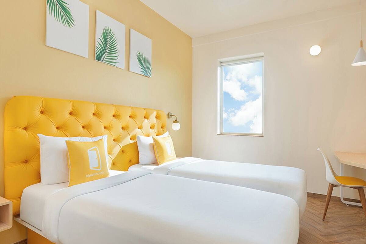 Une chambre d’hôtel avec une tête de lit jaune à motifs capitonnés, deux lits, un bureau et une fenêtre donnant sur le ciel.