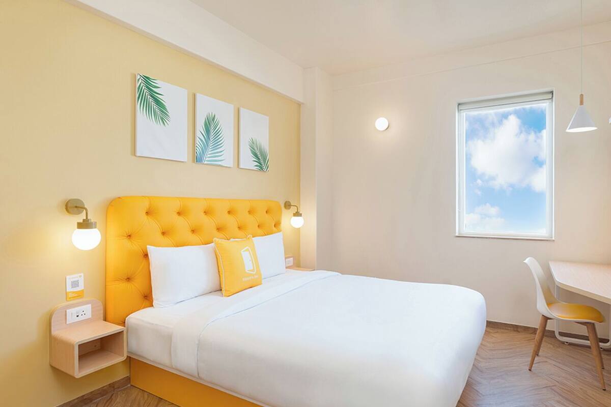 Une chambre avec un grand lit, un bureau et une chaise. Il y a des lampes fixées au mur et une fenêtre donnant sur le ciel.