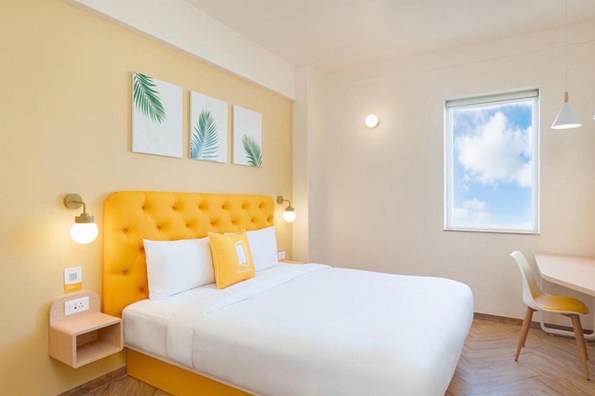 A bedroom with a yellow tufted headboard, white bedding, a small bedside table, a desk with a chair, and a window with a view of the sky.