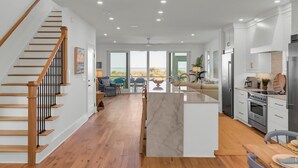 A spacious kitchen with a central island, wooden staircase, and a view of the outdoors. - New Construction 5BR | Designer Coastal Home in Wild Dunes (Isle of Palms)