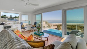 A spacious living area with a large window overlooking the beach, a ceiling fan, and a view of the ocean. - New Construction 5BR | Designer Coastal Home in Wild Dunes (Isle of Palms)