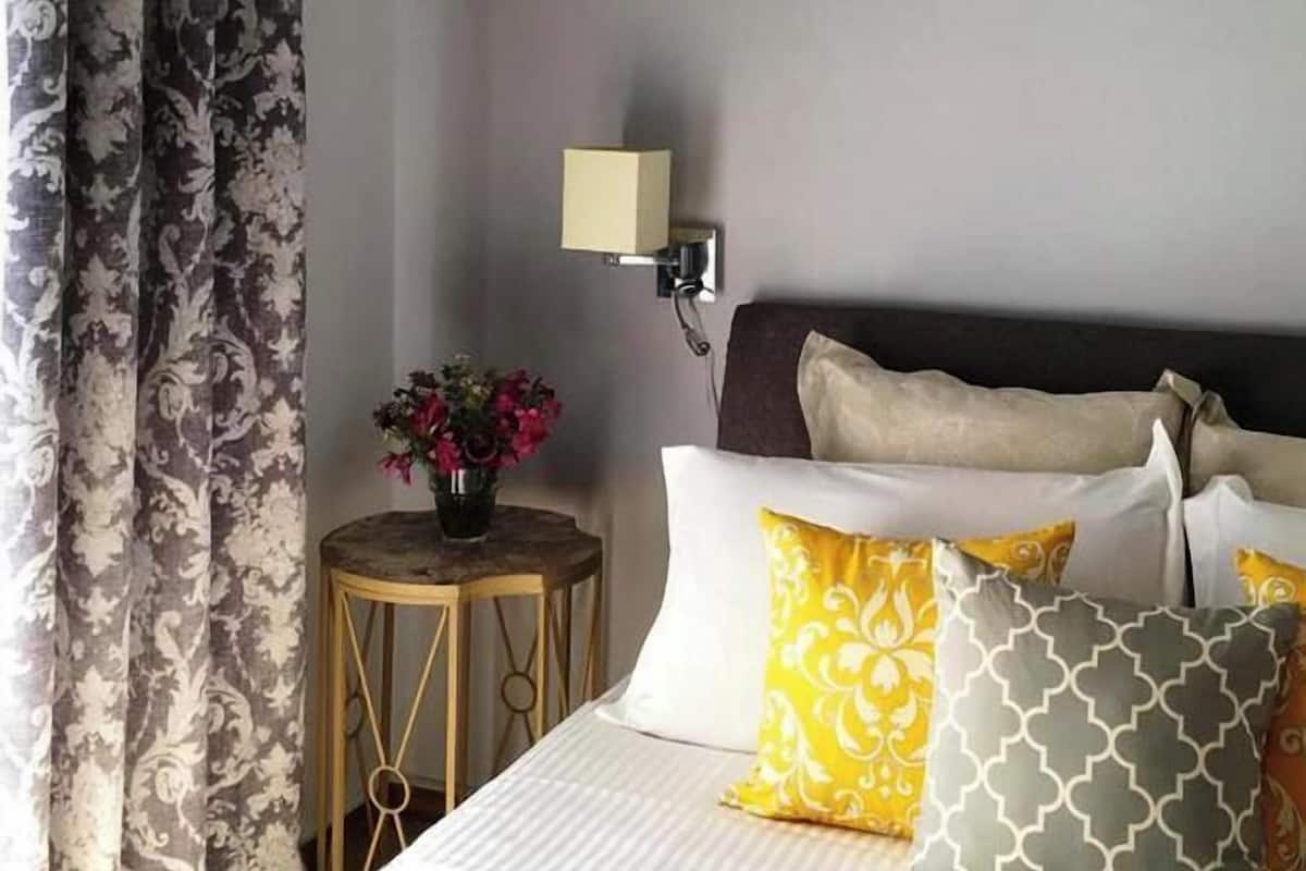 Ein ordentlich bezogenes Bett mit mehreren Kissen, ein Nachttisch mit Blumenvase und eine Wandlampe.