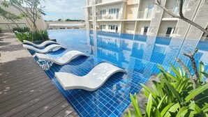 Une piscine aux carreaux bleus et aux chaises longues blanches, entourée de verdure et bordée d’un immeuble à plusieurs étages.