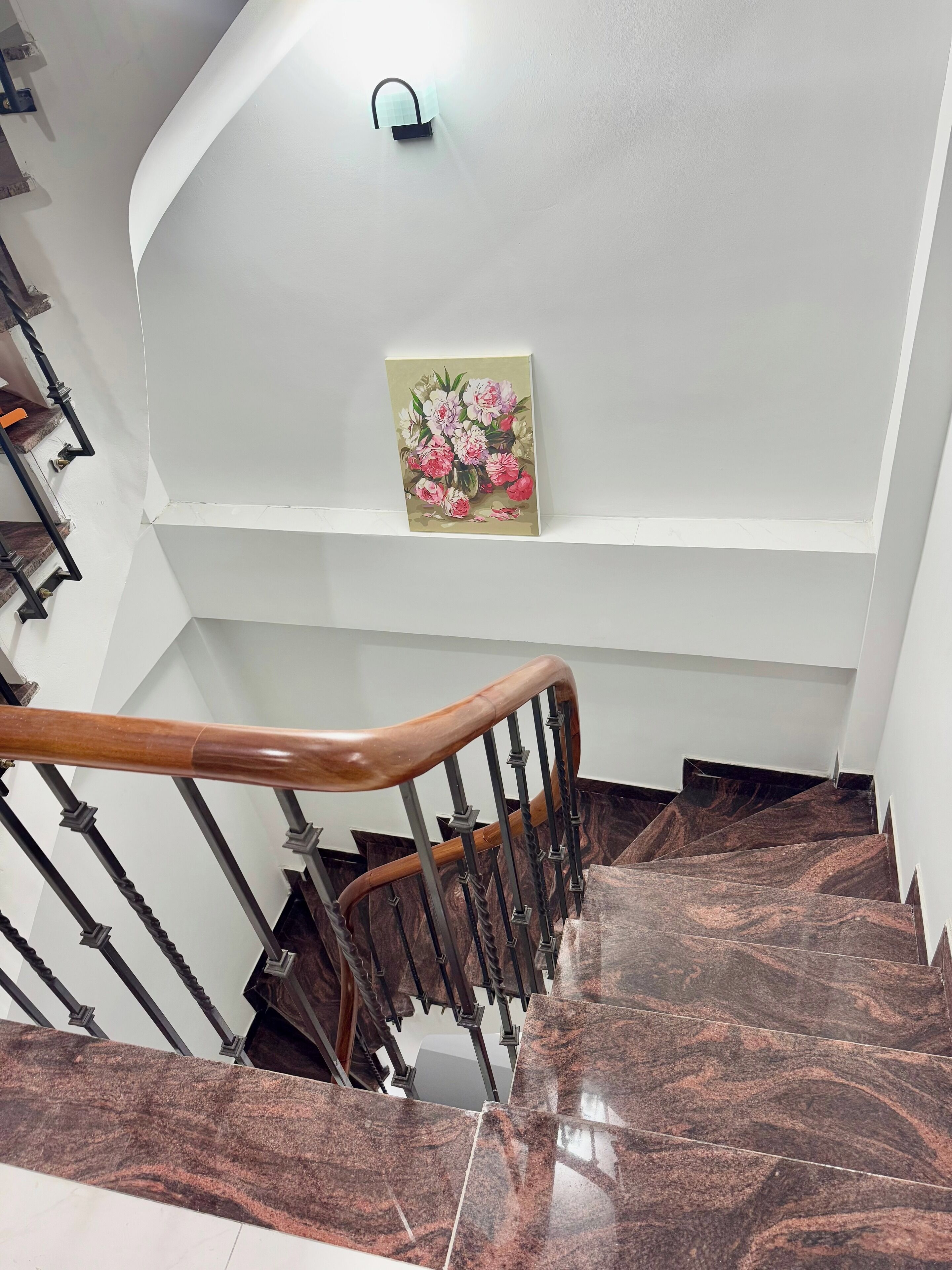 A staircase with marble steps and wooden handrails, leading to a landing with a wall-mounted light fixture and a framed floral artwork.