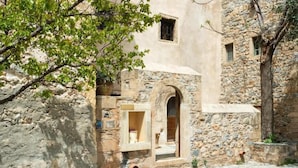 Un bâtiment en pierre avec une petite porte d’entrée en arc de cercle, une fenêtre et un arbre au premier plan.