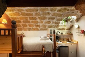 A cozy room with a stone wall, a wooden bed, a small kitchenette, and a window with a view of greenery.