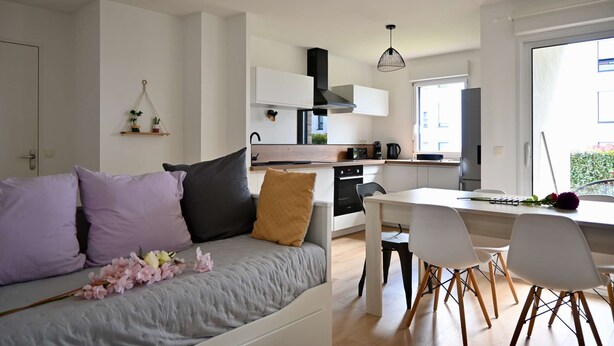 Een moderne, goed verlichte woonkamer met een grijze bank, een witte eettafel en stoelen. Achtergrond toont een keuken met witte kastjes en een zwarte afzuigkap.