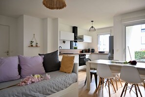 A modern, well-lit living room with a grey sofa, white dining table, and chairs. A kitchen with white cabinets and a black range hood is visible in the background. - Spacious, modern apartment 10 minutes from the port of Vannes (Vannes)