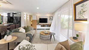 Una sala de estar moderna con sofá, mesa de centro y televisor. Hay una zona de cocina con refrigerador y estufa.