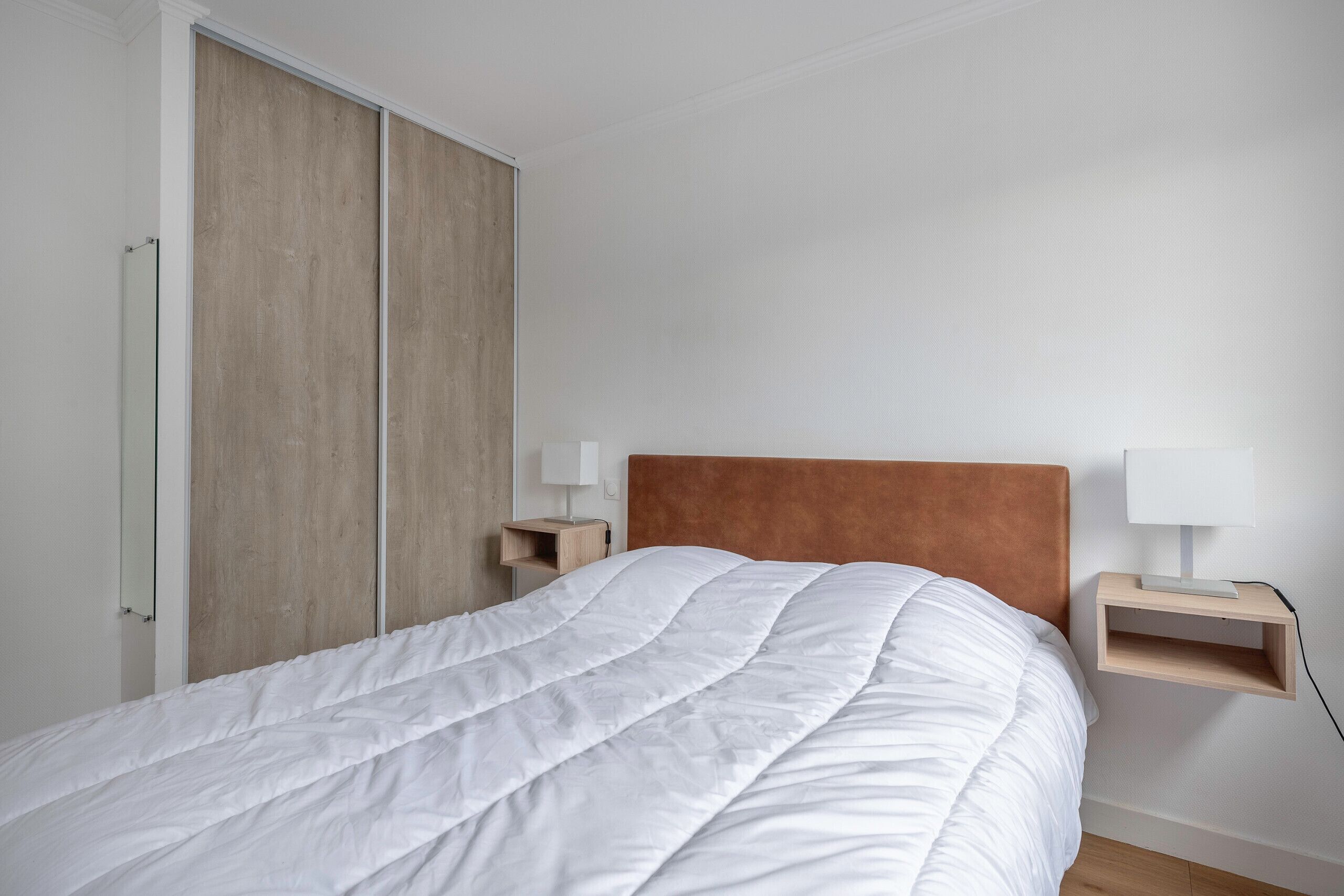A bedroom with a large bed, wooden wardrobe, bedside tables with lamps, and a plain white wall.