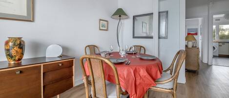 Un comedor con una mesa redonda cubierta con un mantel rojo, rodeada de sillas de mimbre. A la izquierda, un aparador de madera con cajones, y sobre él, un jarrón con flores.
