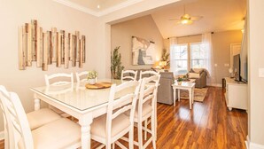 A dining area with a glass-top table and white chairs, a living room with a sofa and a TV, and a ceiling fan.