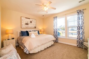 Ein Schlafzimmer mit Bett, Nachttischen, Deckenventilator, einem Fenster mit Vorhängen und einem Wandbild.