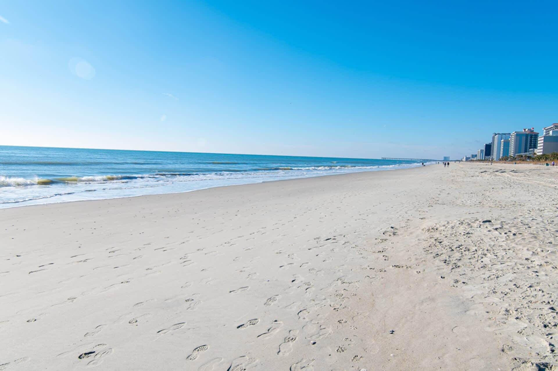 A wide sandy beach with footprints leading to the water, a clear blue sky, and buildings in the background.