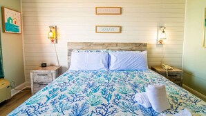 A bedroom with a bed, two bedside tables, a nightstand, a wall-mounted light, and two signs on the wall. - Oceanfront Pet Friendly Modern Suite (Myrtle Beach)