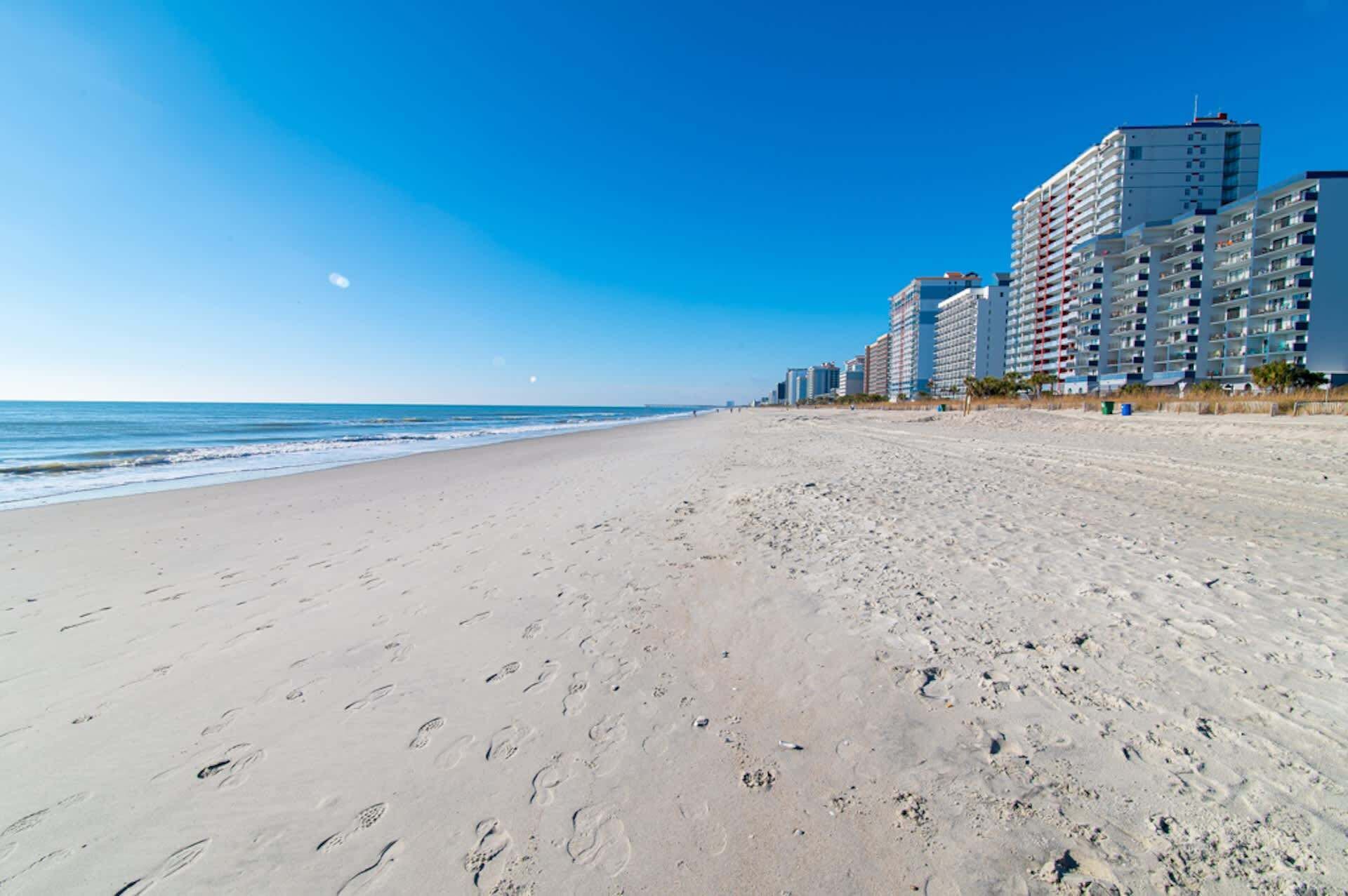 A wide sandy beach with footprints, a clear blue sky, and high-rise buildings in the background.