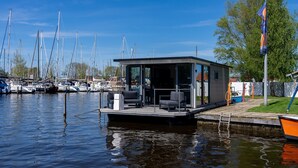 A floating structure with a covered seating area and a small kitchen, docked at a marina with multiple boats. - Houseboat in Heeg With Stunning Marina Views (Heeg)