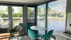 A modern room with a dining area, a kitchen, and a view of a body of water through large windows. - Houseboat in Heeg With Stunning Marina Views (Heeg)