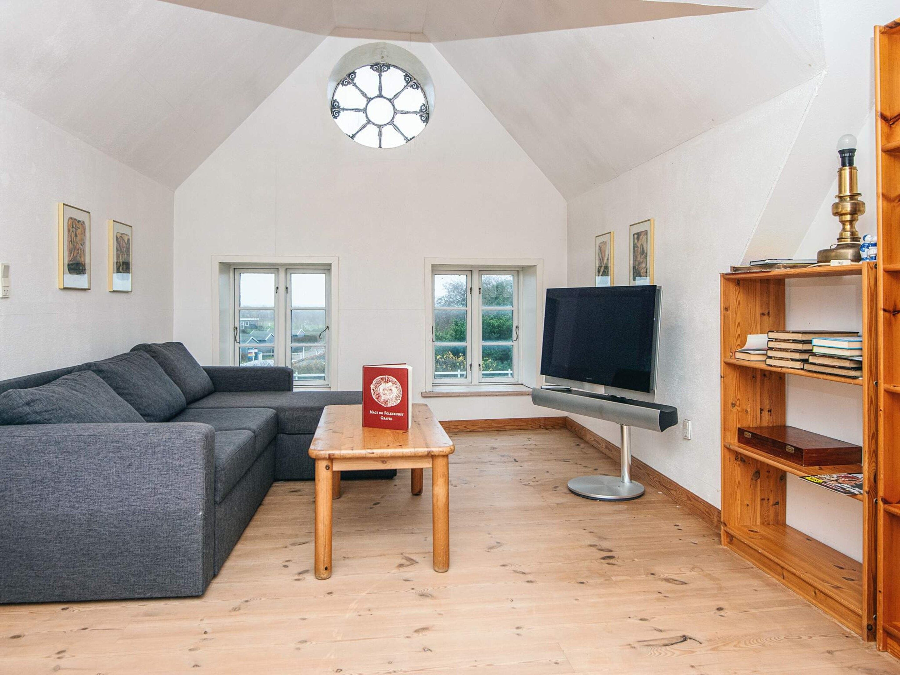 A living room with a grey sofa, a wooden coffee table, a flat-screen TV, a wooden bookshelf, and a window with a view of greenery.