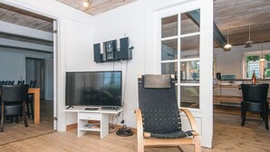 Uma sala de estar moderna com uma televisão de tela plana grande, uma poltrona de madeira e um espaço de jantar com mesa e cadeiras.
