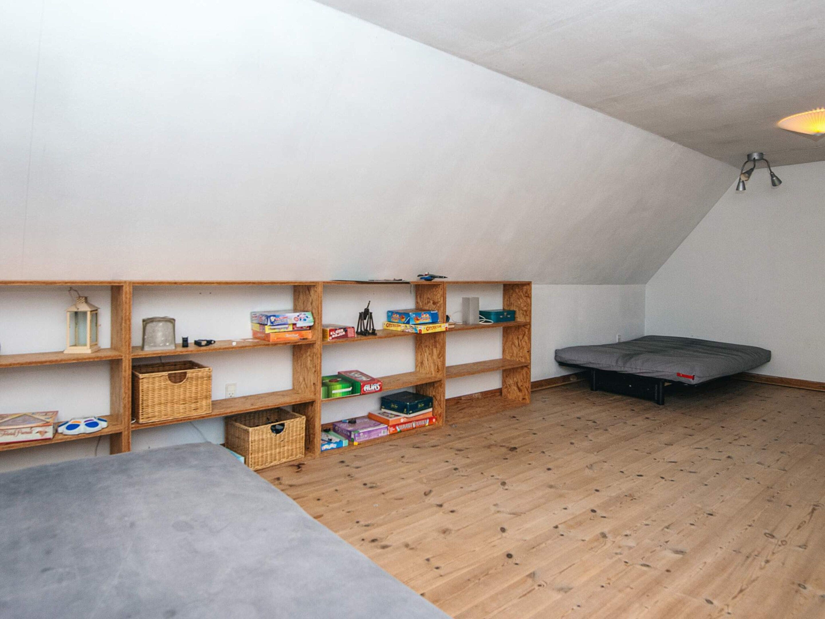 Ein Zimmer mit schrägem Dach, Holzregalen mit verschiedenen Gegenständen, einer Matratze und einem Betonzementboden.