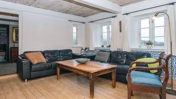 Una sala de estar con una mesa de centro de madera, sofás de cuero negro, un sillón tapizado en azul y vistas al exterior a través de ventanas.