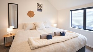 A neatly arranged bedroom with a large bed, two bedside lamps, a wall mirror, a framed artwork, and a window with a view of buildings. - Newly built unit in St Kilda. Modern amenities (Dunedin)